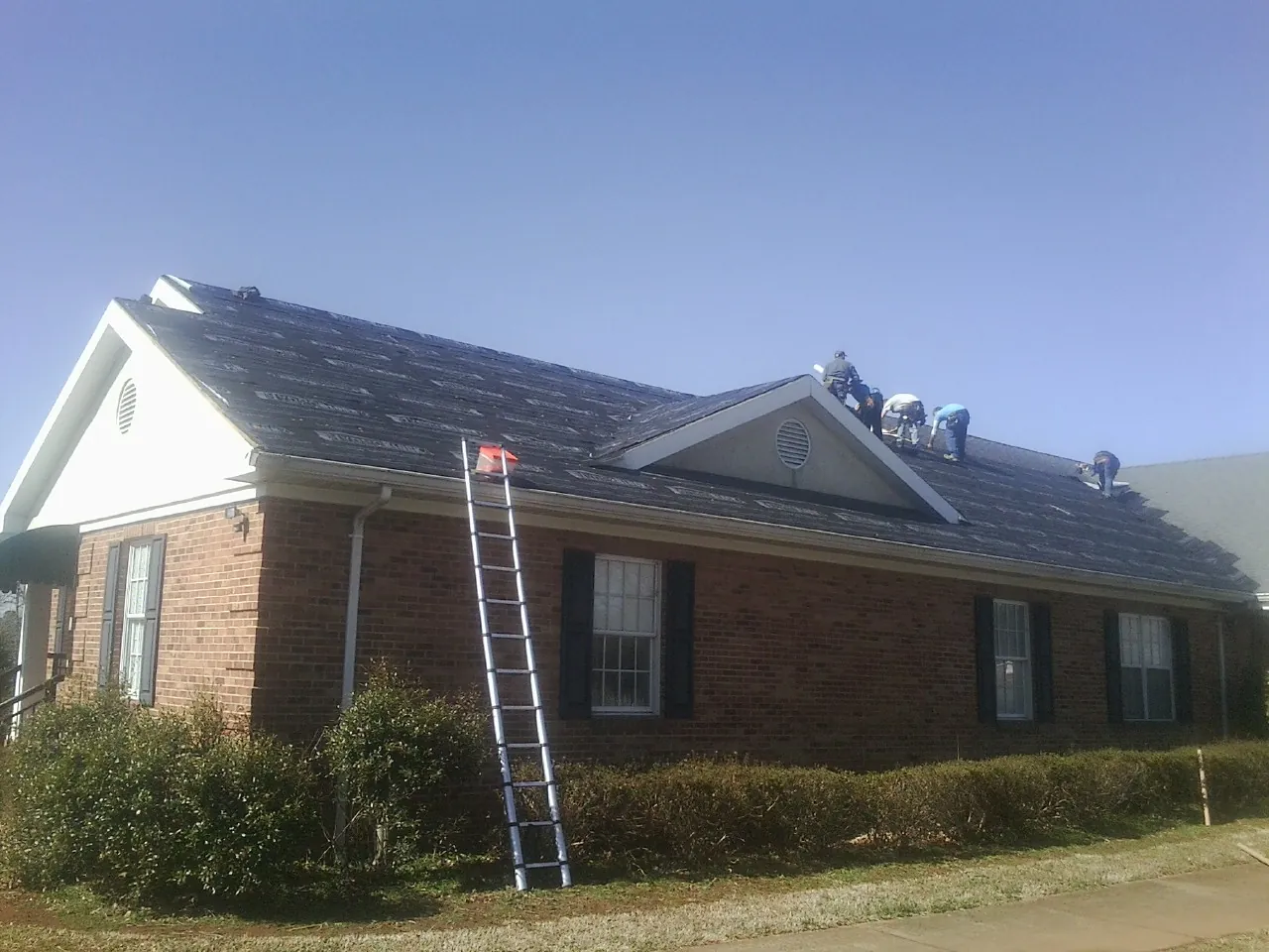 Crew tearing shingles off a brick ranch roof with a ladder set — Metal Roof Repair work by our crew in Sunman