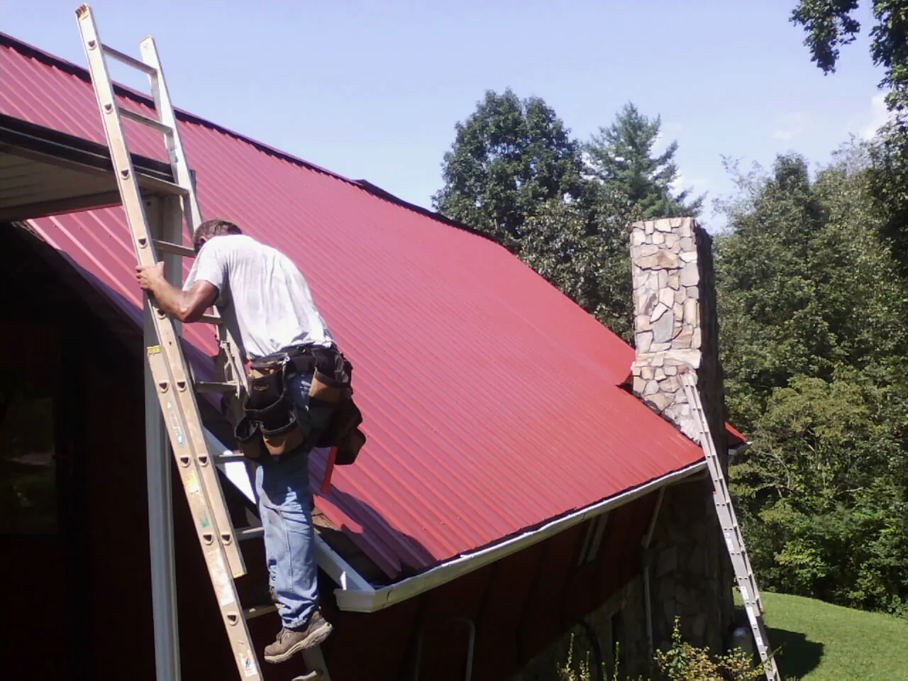 Roofer on a ladder beside a red metal roof and stone chimney — Soffit & Fascia Repair by Roofing Brothers in Sunman